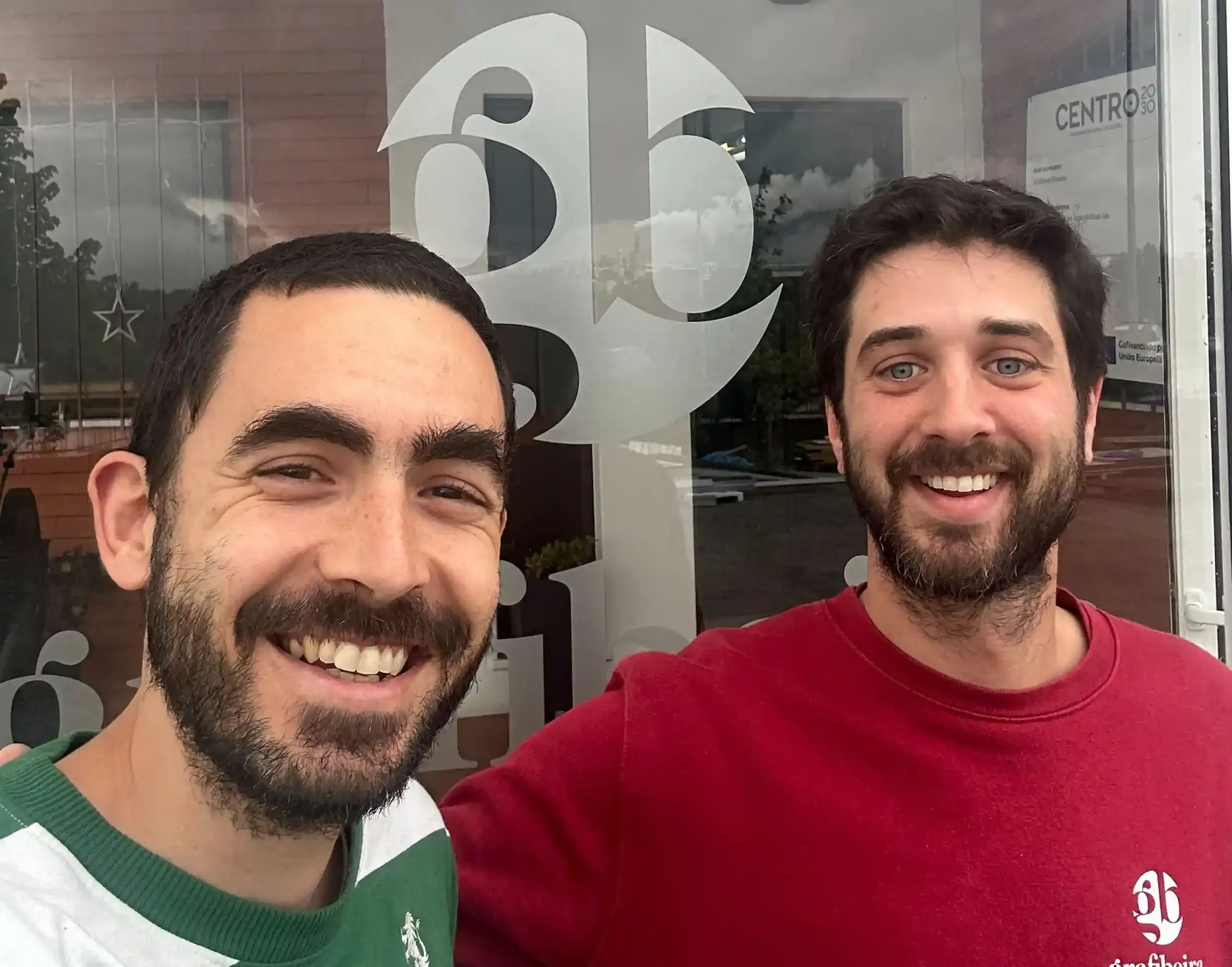 Two smiling men in front of a window with the Grafibeira logo, one wearing a red Grafibeira sweatshirt